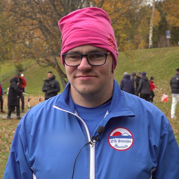 Henrik Berglund, från orienteringsklubben OK Storsjön, står utomhus med ljusblå klubbjacka och rosa mössa.