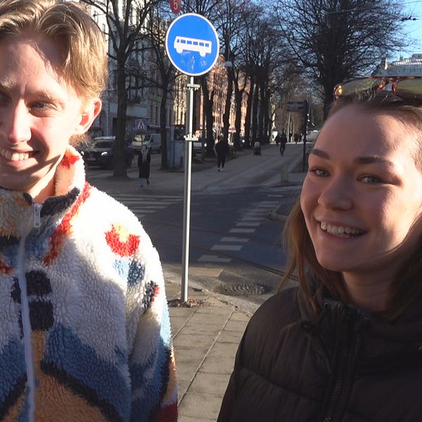 Två personer står på en trottoar i en stadsmiljö. En blå skylt, som indikerar en busshållplats, är synlig i bakgrunden. En bred gata kantad av byggnader och träd sträcker sig bortom personerna. Det är en solig dag.