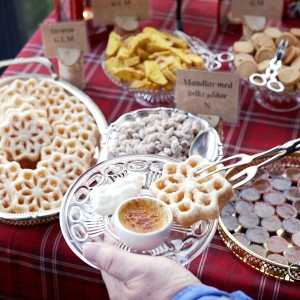 En hand plockar upp en krispig kaka från en silverfärgad tallrik, där det även finns en liten skål med en krämig dip. I bakgrunden syns flera olika sorters godsaker som är vackert presenterade på ett rödvitrutigt bord. Små skyltar beskriver de olika delikatesserna.