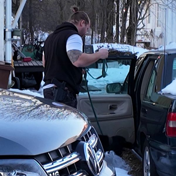 En polis står vid en bil som har en öppen dörr med en hund i koppel, I bakgrunden syns en byggnad och en annan bil.