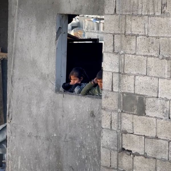 Två barn kikar ut genom ett fönster i en betongbyggnad i Gaza.
