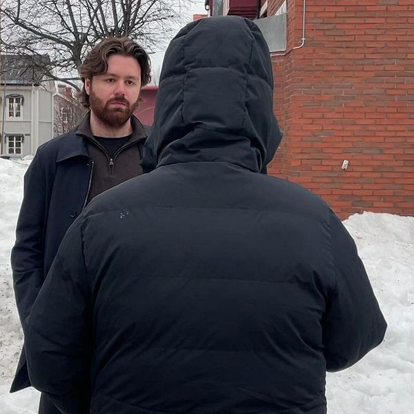 En anonym person som har på sig en svart jacka med luva. Mittemot står en reporter – en man med brunt hår