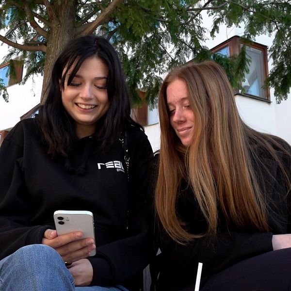 En kvinna tittar på sin smartphone medan hon sitter bredvid en annan kvinna utomhus. De är omgivna av träd och en byggnad med fönster i bakgrunden. Det ser ut som en avslappnad stund i en park eller på en uteservering.