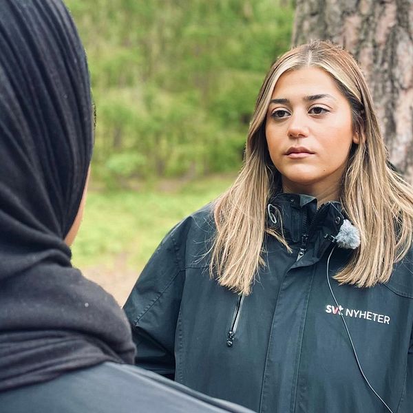 Två personer står vid en trädstam i en grönskande miljö, med fokus på en intervju. Den ena personen bär en svart klädsel, medan den andra har långt, ljust hår och en svart jacka med en logotyp på bröstet. Den omgivande naturen ger en lugn bakgrund för samtalet.