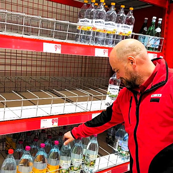 En person i en röd jacka står vid en hylla i en matbutik och inspekterar flaskor med drycker. Hyllan är delvis tom, med några flaskor av olika sorter, inklusive vatten och läsk. Prisskyltar är synliga ovanför hyllorna.