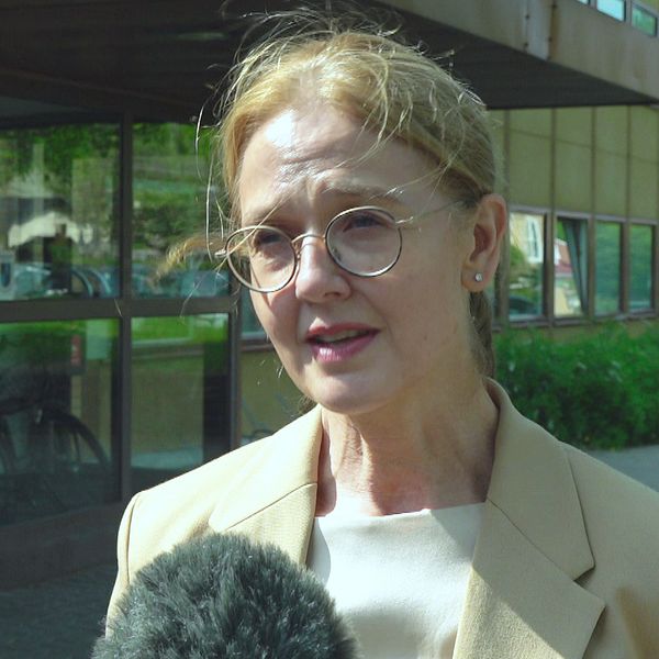En kvinna i beige kavaj står framför en gul byggnad