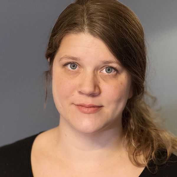 En kvinna med mörkt blont hår uppsatt i en låg hästsvans syns. Hon bär en svart tröja. Bakgrunden är en oskarp grå vägg.