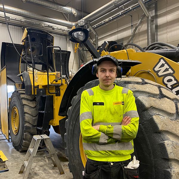 En stor gul entreprenadmaskin inne i en verkstad med arbetare i varselkläder.