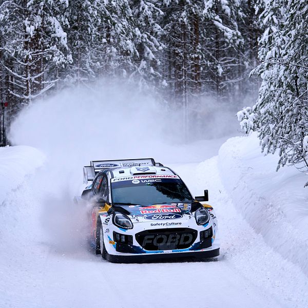 En rallybil kör på en snötäckt väg omgiven av tallar, med snö som sprutar upp bakom bilen.
