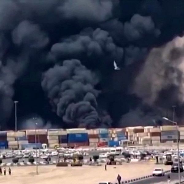 Tjock svart rök stiger från brand vid en containerhamn, bilar och folk i förgrunden.