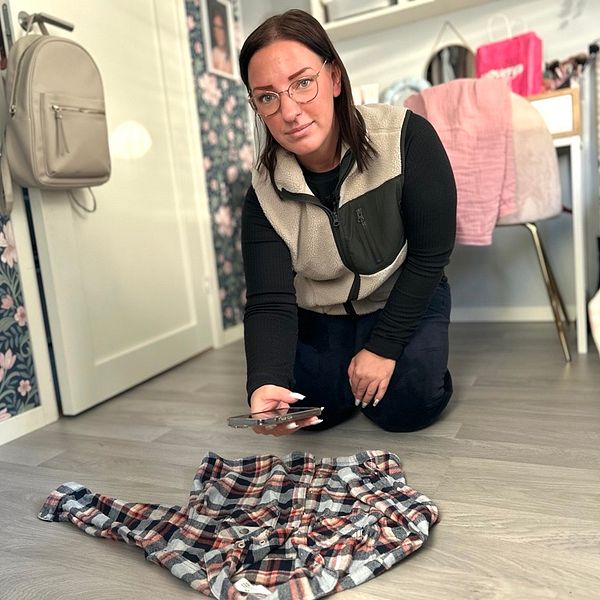En person knäböjer på golvet i ett rum med blommönstrad tapet och fotograferar en rutig skjorta som ligger på golvet. I bakgrunden syns en ryggsäck och rosa textilier.