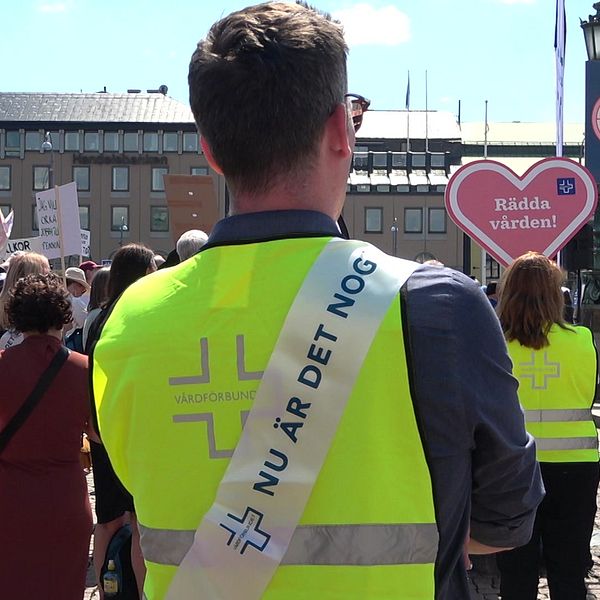 Flera personer strejkar på ett torg. En man med gul väst från Vårdförbundet har ett budskap på ryggen med texten ”nu är det nog”.