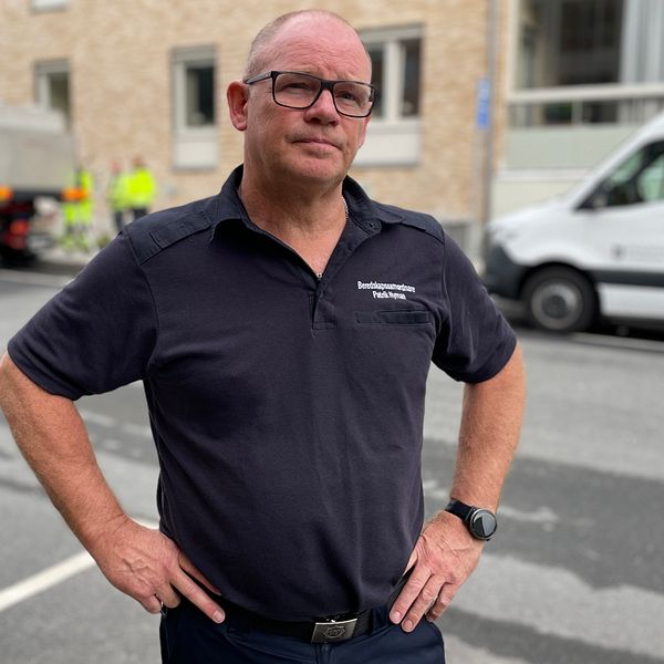 Patrik Nyman beredskapssamordnare i Jönköpings kommun står med händerna på höfterna på en gata, iförd en mörkblå arbetsuniform med broderad text. I bakgrunden syns en lastbil och annat arbete som pågår, vilket indikerar en aktivitet relaterad till bygg eller underhåll. Solen skiner och det är en klar dag.