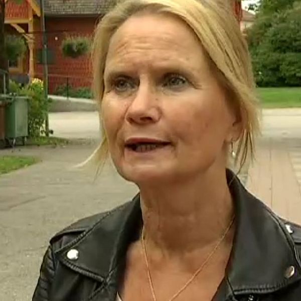 Kvinna i svart läderjacka står på en parkväg, omgiven av grönska och byggnader i bakgrunden. Det är en klar dag och en parkstruktur syns i avståndet.