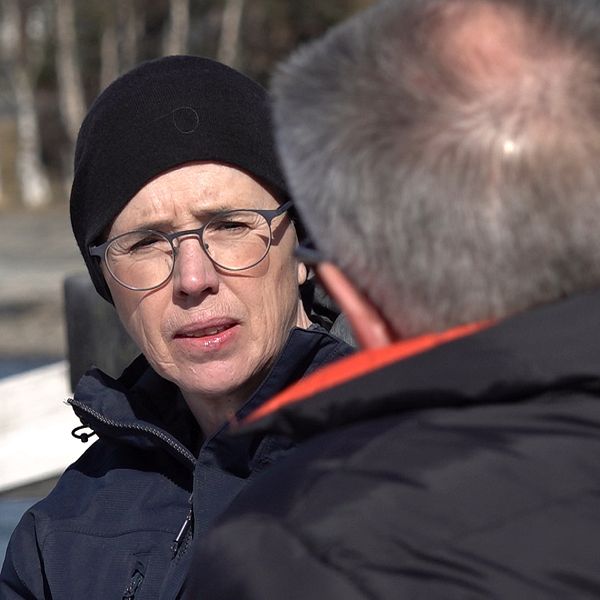 Två personer samtalar i en utomhusmiljö med snö och träd i bakgrunden.
