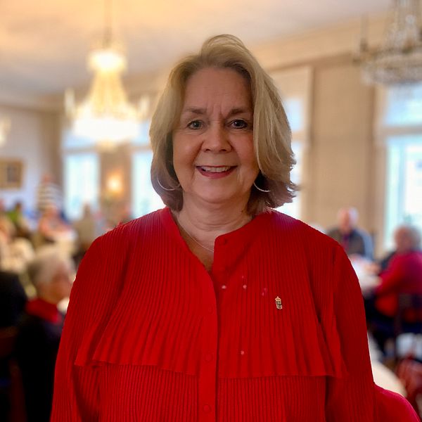 En person i en röd blus står i förgrunden av en social samling, där många andra personer är synliga i bakgrunden i en festlig miljö. Rummet har ljusa väggar och är inrett med kristallkronor och fönster som släpper in naturligt ljus. De andra deltagarna verkar vara engagerade i samtal eller aktiviteter.