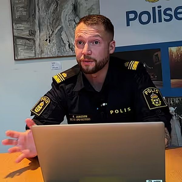 En polis, klädd i uniform, sitter vid ett bord framför en laptop. Bakgrunden visar en vägg med en stor skylt med texten ”Polisen” och en bild som relaterar till polisarbetet. Polisen gestikulerar med handen, vilket antyder att han diskuterar något viktigt.