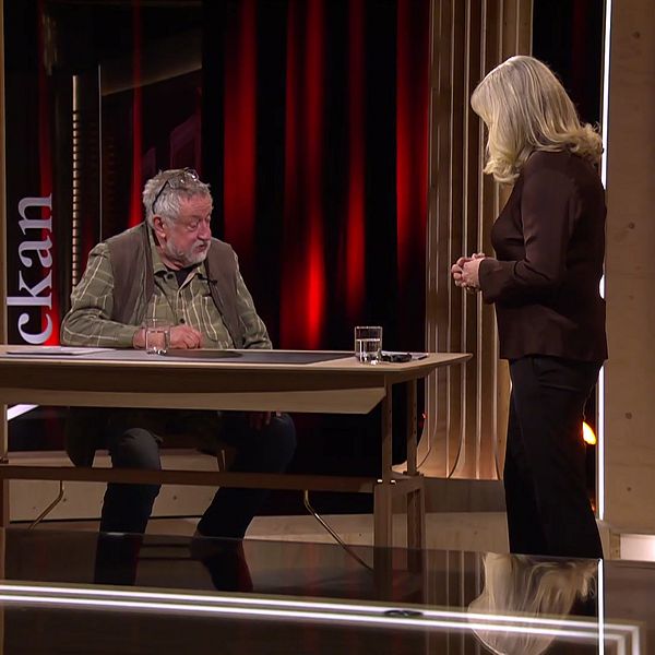 Två personer vid ett bord i en TV-studio för ”Veckan”. Camilla Kvartoft står upp, Leif GW Persson sitter.