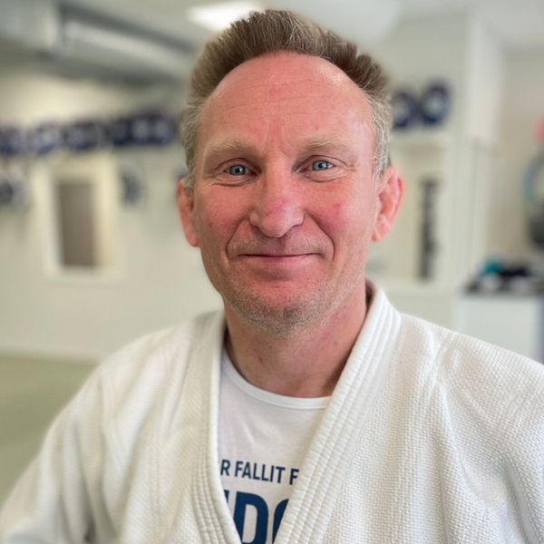 En person i judodräkt står i en träningslokal med pokaler i bakgrunden.