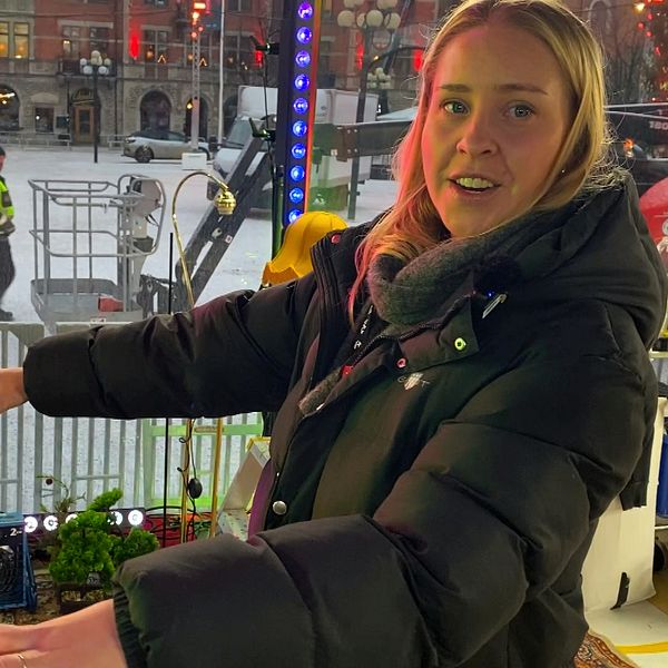 En person står i Musikhjälpens bur och håller ut armarna. Bakom personen syns en man i uniform som arbetar i området, samt en lyftplattform och en stängselstruktur. Den omgivande staden har lampor som lyser och skapar en livlig atmosfär.