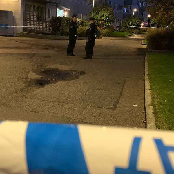 ALT-text: Två poliser står i en mörk bostadsområde där avspärrningar med polisband syns i förgrunden. En eventuell brottsplats är markerad i gatan.