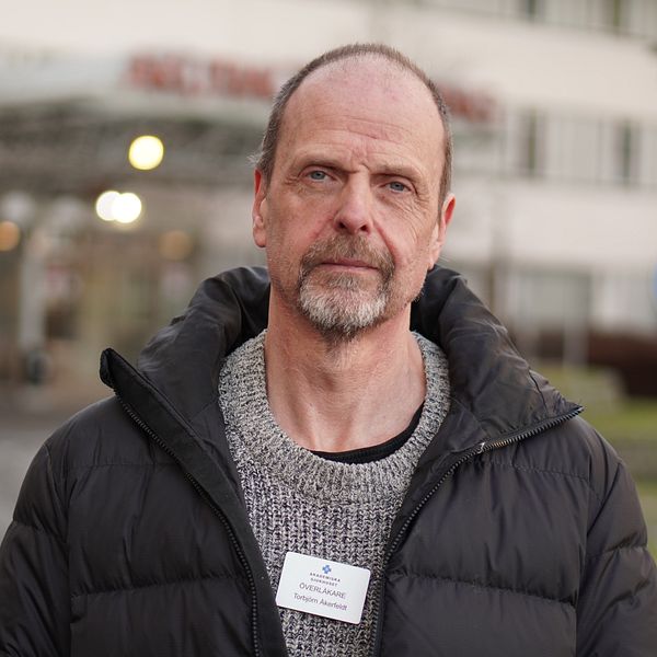 En person i en svart jacka och en grå tröja står framför en byggnad med en skylt. Personen bär ett namnskylt på bröstet. I bakgrunden syns en parkeringsskylt och träd.
