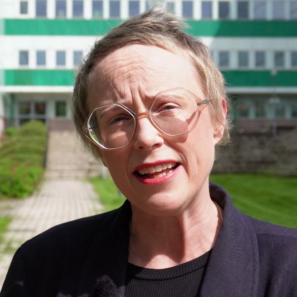 Kvinna med ljust kort hår står framför en vit byggnad med gröna detaljer. Hon har glasögon och mörk kavaj.