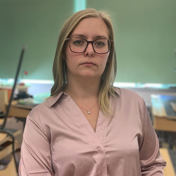 Kvinna i en rosa skjorta står framför en klassrumsmiljö med bord och stolar i bakgrunden. Ljusa fönster ger naturligt ljus till rummet. Lärare, lärarbehörighet, skola, varför få behöriga lärare?