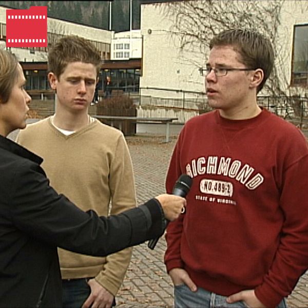 En journalist intervjuar två unga män framför en byggnad med parkeringsplatser och träd omkring. Den ena mannen bär en beige tröja och den andra en röd tröja med texten ”Richmond”.