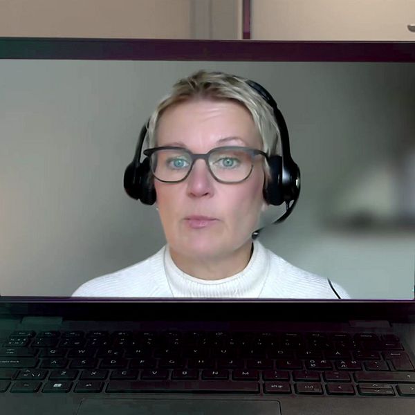 En laptop står på ett bord och visar ett videosamtal. Skärmen visar en person som bär hörlurar och en vit tröja, medan bakgrunden är oskarp.