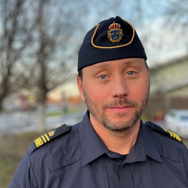 En man i uniform med en militärisk kap över huvudet står framför en suddig bakgrund med träd och byggnader. Han ser fokuserad ut och är klädd i en mörkblå skjorta med guldiga insignier på axlarna.