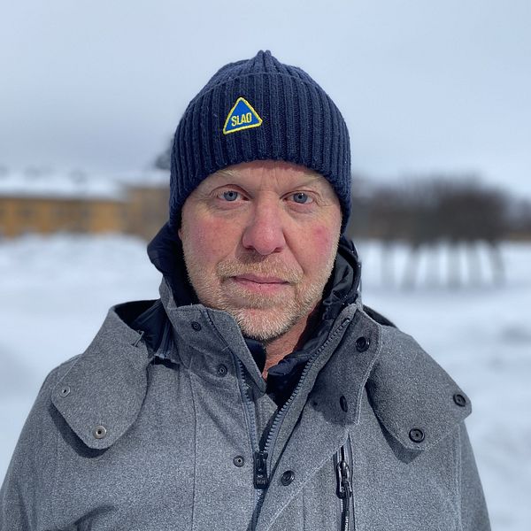 Slao:s vd Magnus Noppa, klädd i en grå vinterjacka och mörk mössa står utomhus på en snöig plats, med en suddig byggnad och träd i bakgrunden.