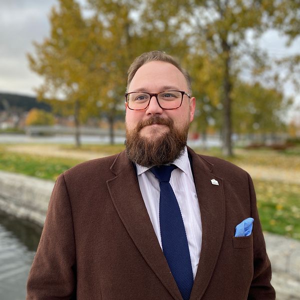 Jonas Andersson (S) med glaögon, brunt skägg, brun kavaj, vit skjorta och blå slips står vid ett vatten.