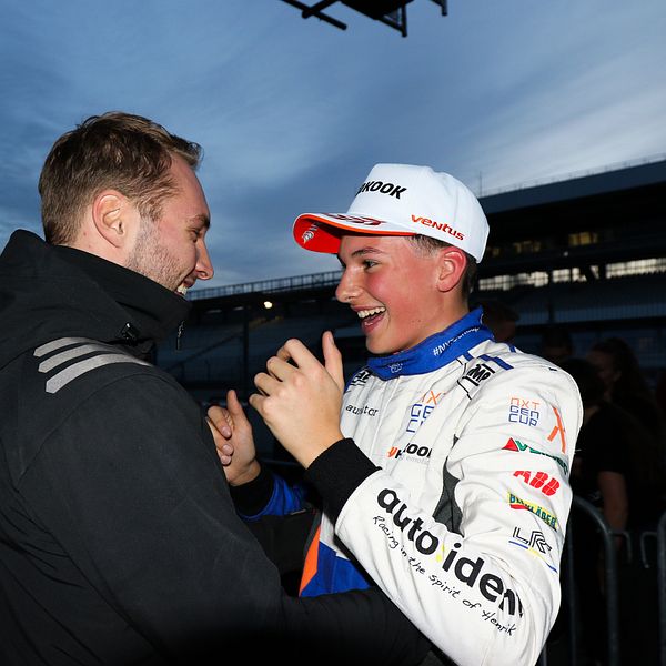 Två personer interagerar i en pitområde vid en racinghändelse. Den ena personen i en mörk jacka står nära en annan person iförd en motorsportoverall med sponsorlogotyper. Bakgrunden visar en racingbil och en dimmig himmel som antyder skymning.