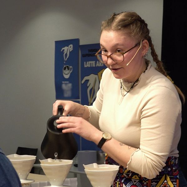 En kvinna häller kaffe från en kanna i vita filter på ett bord medan en man med jeansjacka tittar på, i vad som verkar vara ett kaffeevenemang eller en kaffedemonstration.