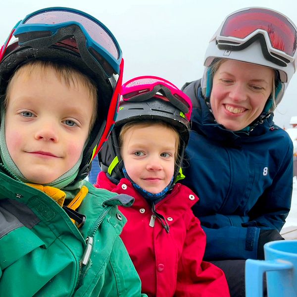 Två yngre barn och en mamma i skidkläder och hjälmar frmför en skidbacke