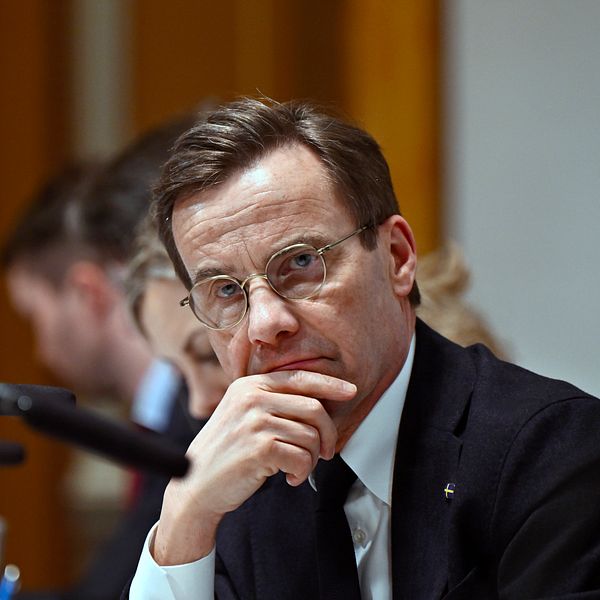 Statsminister Ulf Kristersson i kostym lyssnar under ett möte med mikrofoner och suddiga personer i bakgrunden.
