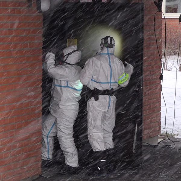 Två personer – poliser – klädda i skyddsutrustning arbetar vid en ingång till en byggnad, ett soprum, i snöigt väder. En av dem håller upp en lampa, vilket skapar en ljuspunkt i det mörka utrymmet inuti. I bakgrunden syns en soptunna och snö på marken.