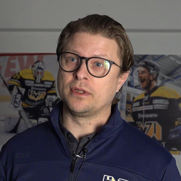 Hockeytränare med blå jacka framför tre matchfotografier på väggen.