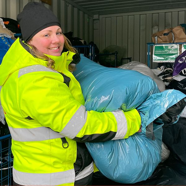 Sarah Törnqvist, ansvarig på återvinningscentralerna i Landskrona och Svalöv, bär på en stor blå plastpåse fylld med kläder. Hon står inne i en stor container fylld av textilier både i plastpåsar och som ligger löst. Bakgrunden visar kaos av kläder och kassar som lagrats.