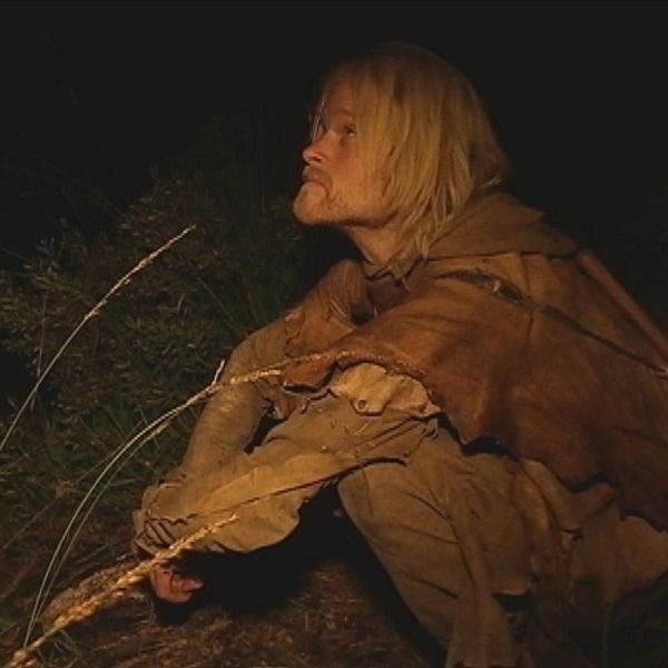 Person i bruna kläder hukar i en mörk skogsmiljö.