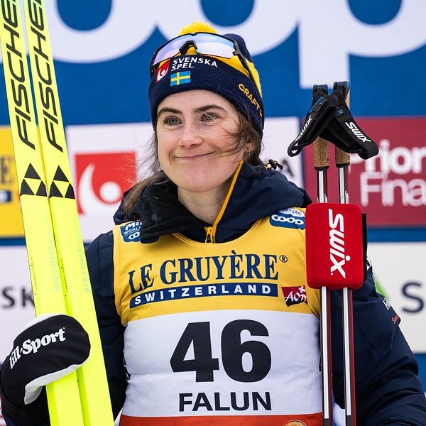 En skidåkare klädd i tävlingsuniform med nummer 46 håller sina skidor och stavar vid sidan av en bakgrund med sponsorer i Falun.