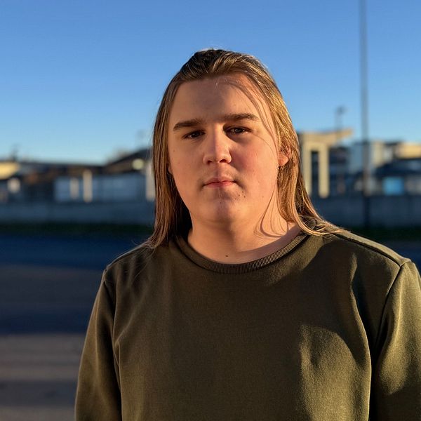 En person står framför en buss på ett resecentrum, omgiven av en klarblå himmel och tågrälsen i bakgrunden. Personen är klädd i en grön tröja och blicken är riktad mot kameran. Bussens färgschema inkluderar röd och vit.