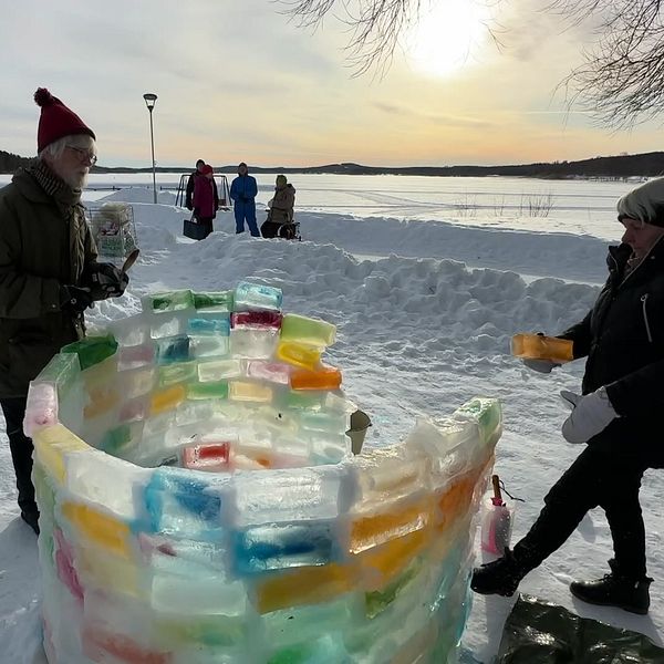 Två personer bygger en färgglad igloo på en snötäckt plats vid en sjö i solnedgången.