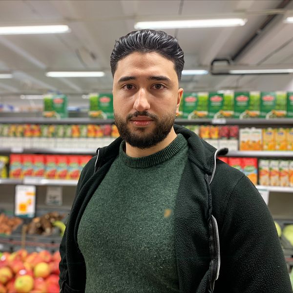 En man står i en matbutik och tittar rakt in i kameran. Grönsaker och andra varor i bakgrunden.