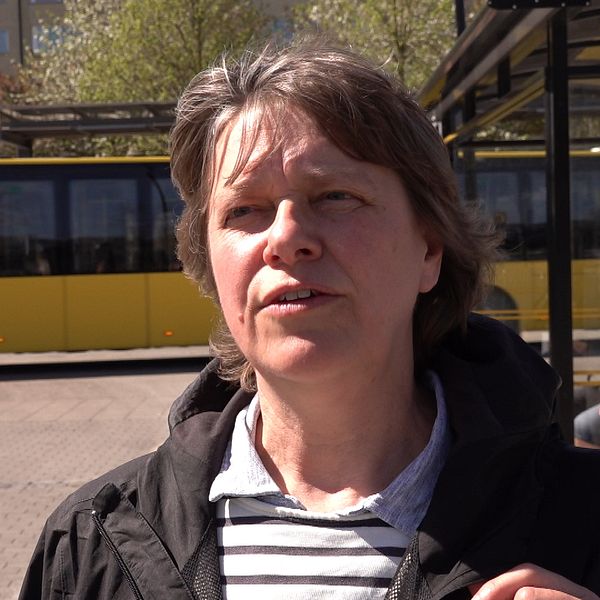 En kvinna står vid en busshållplats och en gul buss syns i bakgrunden.