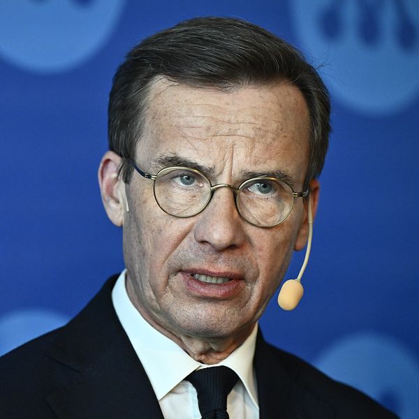 Statsminister Ulf Kristersson i kostym framför blå bakgrund med Moderaternas logo.