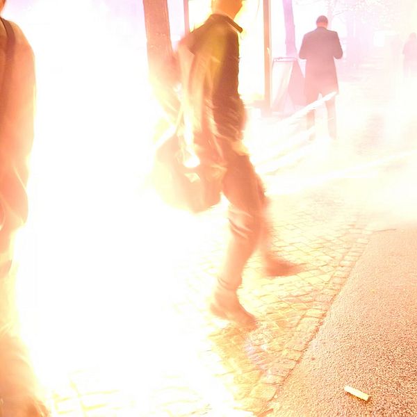 Personer som skyndar sig förbi en explosion eller kraftig ljuseffekt i en stadsmiljö, med rök och ljus som sprider sig i luften. Många verkar vara på väg bort från händelsen, vilket skapar en känsla av hastighet och oro.
