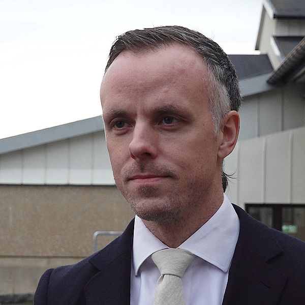 Åklagare David Kahl står utomhus klädd i kostym med en vit skjorta och slips, med en byggnad i bakgrunden och träd utan löv i förgrunden som indikerar tidig vinter.