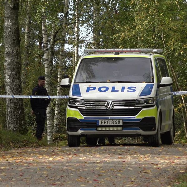 En polisbil står på en grusväg omgiven av träd, med avspärrningsband som markerar en plats för en pågående utredning. En polis fungerar som vakt vid avspärrningen. Höstlöv ligger på marken.